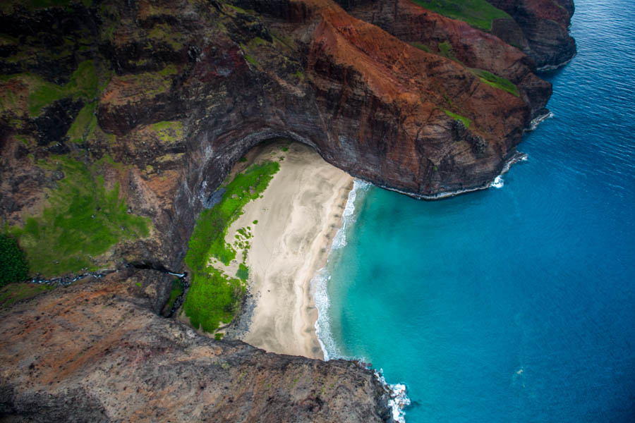 A beach set into a cliff cove seen from the air, turquoise shallows against black rock, Kauai
